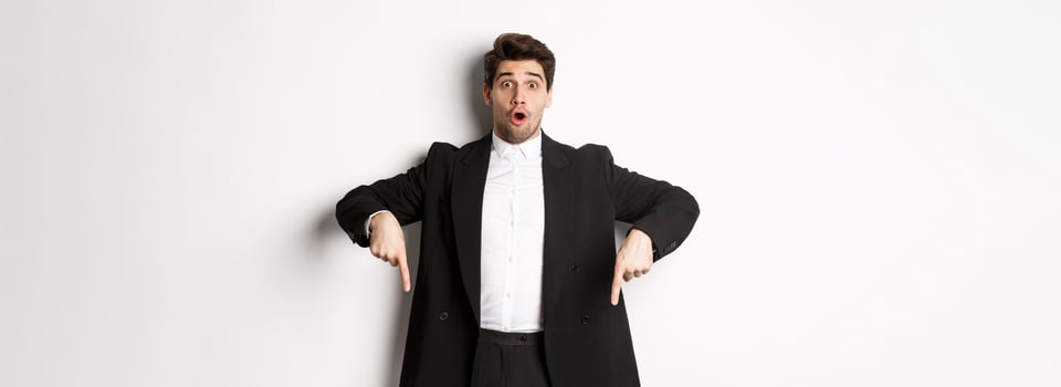 Portrait of surprised and excited man in black suit, pointing fingers down and showing christmas advertisement, standing over white background photo