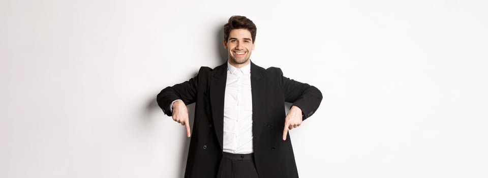 Portrait of good-looking stylish man in black suit, pointing fingers down and smiling, showing winter holidays promo, standing over white background photo