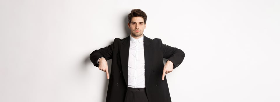 Image of handsome man dressed for formal party, wearing suit and pointing fingers down, showing advertisement or making announcement, standing over white background photo