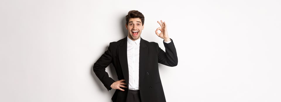 Concept of new year party, celebration and lifestyle. Portrait of pleased handsome guy in black suit, praise something good, showing okay sign in approval, standing over white background photo