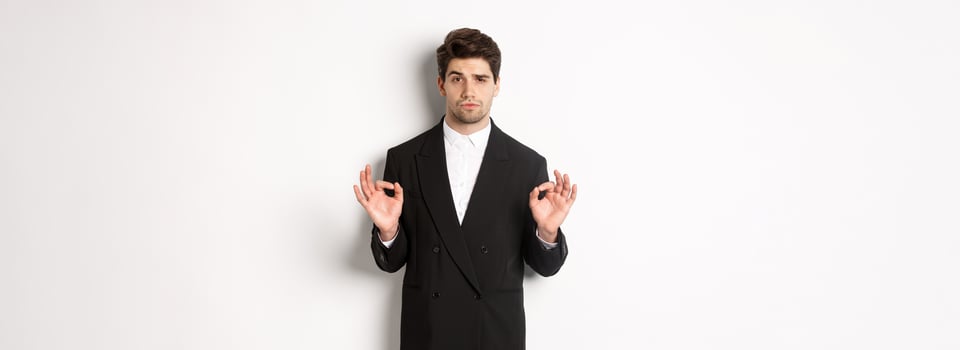 Concept of new year party, celebration and lifestyle. Portrait of confident good-looking man in black suit, showing okay sign and approve something, standing over white background photo