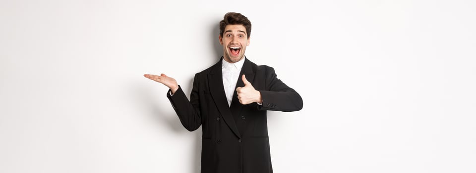 Portrait of hadnsome bearded man in formal suit, showing thumb-up and holding product in hand over white copy space, recommending product, standing over white background photo