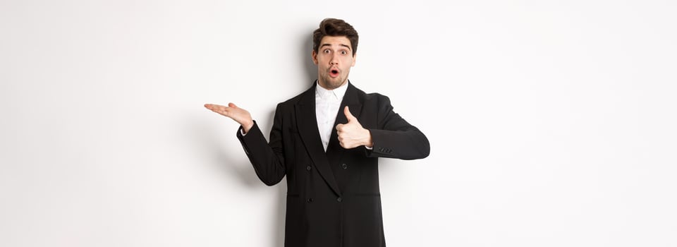 Portrait of hadnsome bearded man in formal suit, showing thumb-up and holding product in hand over white copy space, recommending product, standing over white background photo