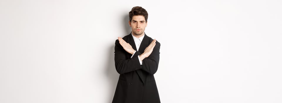 Portrait of serious and concerned man in black suit, showing stop gesture and frowning, making cross to prohibit or decline something, standing over white background photo