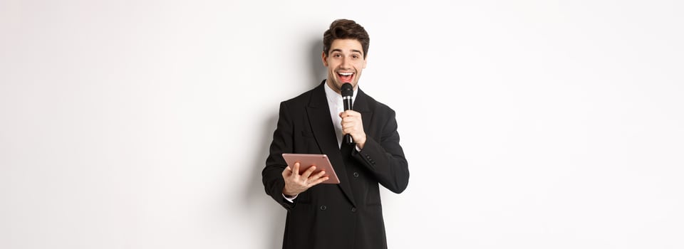Image of handsome male performer, giving a speech, entertain people at party, holding microphone and digital tablet, standing over white background photo