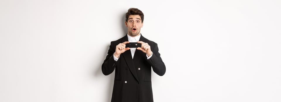 Portrait of handsome businessman in black suit, showing credit card and looking amazed, standing over white background photo