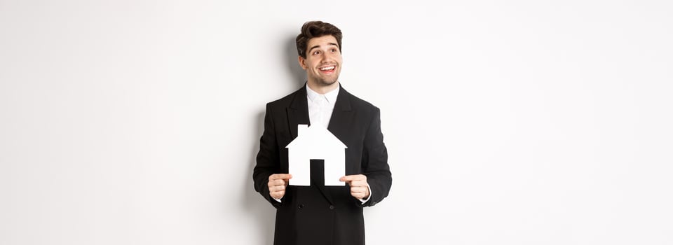 Image of handsome investor in black suit, holding house maket and looking at upper left corner thoughtful, standing against white background photo