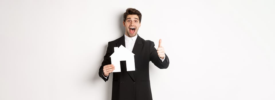 Portrait of excited handsome man looking for a home, showing house and thumb-up, recommending real estate agency, standing against white background photo