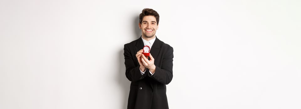 Image of handsome boyfriend in black suit making a proposal, asking to marry him and showing wedding ring, standing over white background photo