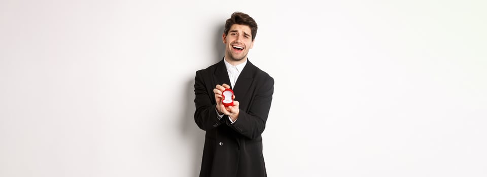Portrait of handsome man in black suit, open box with wedding ring, making a proposal, asking to marry him, standing against white background photo