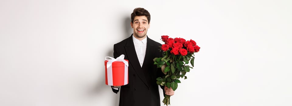 Concept of holidays, relationship and celebration. Image of handsome smiling guy in black suit, holding bouquet of red roses and giving present on new year, white background photo