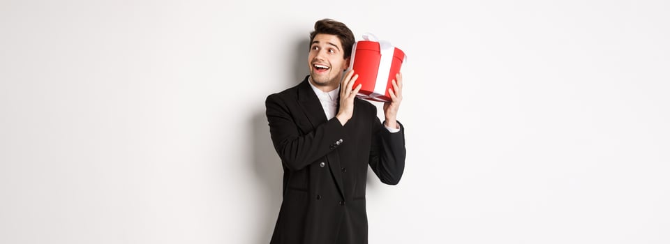 Concept of christmas holidays, celebration and lifestyle. Image of attractive man in black suit, shaking present to guess what inside, enjoying new year, standing against white background photo