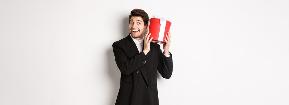 Concept of christmas holidays, celebration and lifestyle. Handsome man in black suit, shaking a box with gift, wondering whats inside, standing against white background photo