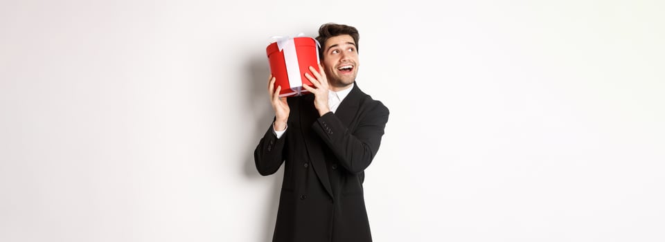 Concept of christmas holidays, celebration and lifestyle. Image of excited man enjoying new year, shaking gift box to guess what inside, standing against white background photo