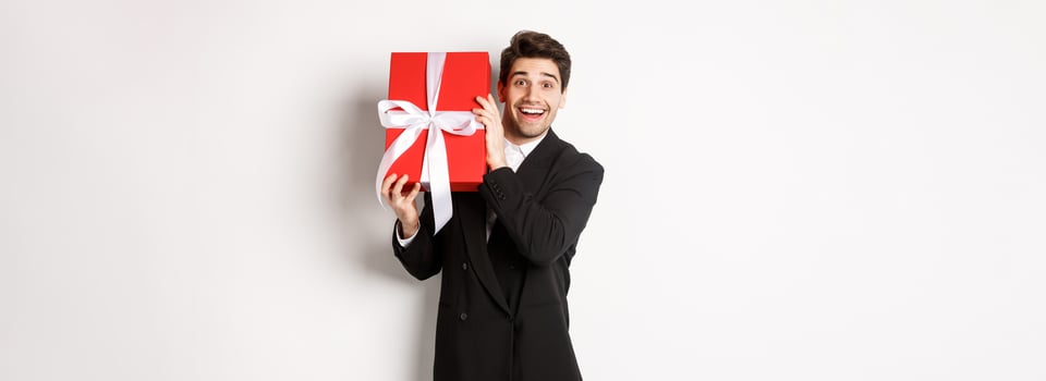 Concept of christmas holidays, celebration and lifestyle. Attractive man in black suit, holding new year gift and smiling, standing with a present over white background photo