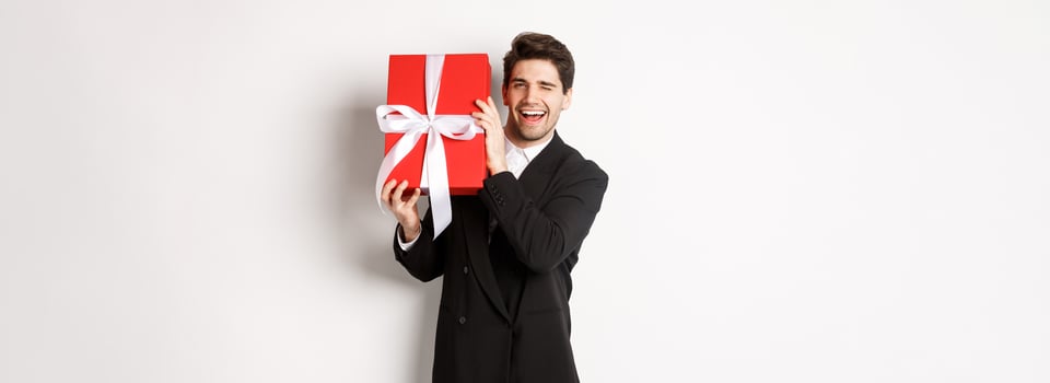 Concept of christmas holidays, celebration and lifestyle. Attractive man in black suit, holding new year gift and smiling, standing with a present over white background photo