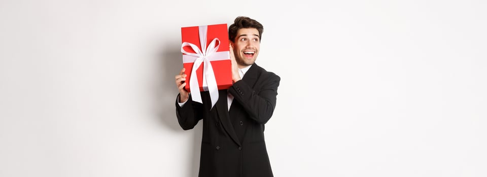 Image of handsome dreamy guy in black suit, shaking box with present to wonder whats inside, standing against white background happy photo
