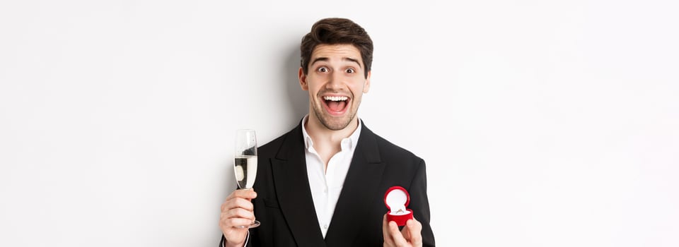 Close-up of handsome man in suit, making a proposal, giving engagement ring and raising glass of champagne, standing against white background photo