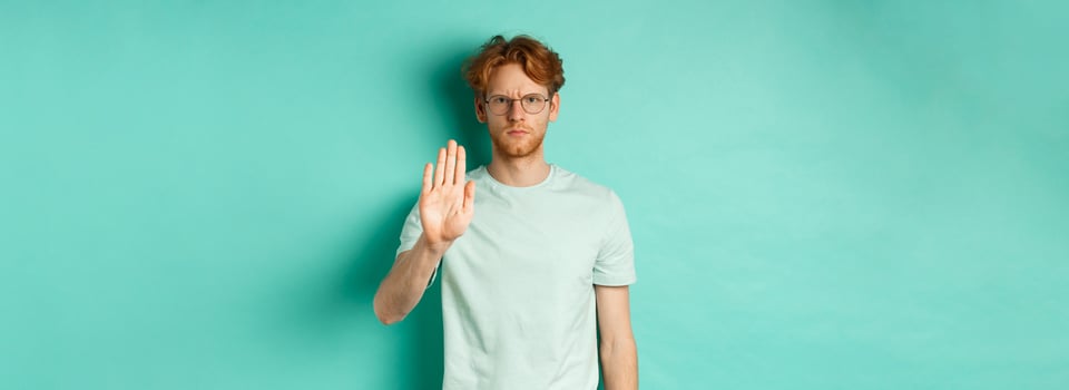 Angry and serious young man with red hair, wearing glasses, showing stop gesture, telling no, disapprove and prohibit something bad, standing over turquoise background photo