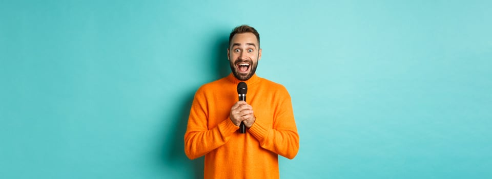 Happy adult man singing karaoke, holding microphone and looking at camera, standing in orange sweater against turquoise background photo