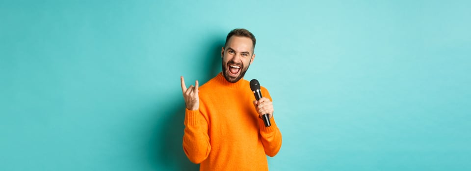 Joyful handsome man singing song in microphone, showing rock-n-roll gesture excited, standing over turquoise background photo