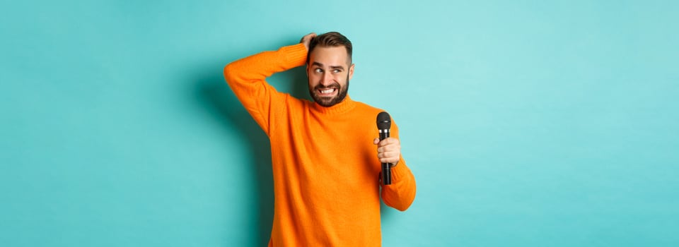 Shy and awkward adult man scratching head, holding microphone before singing, standing over blue background photo