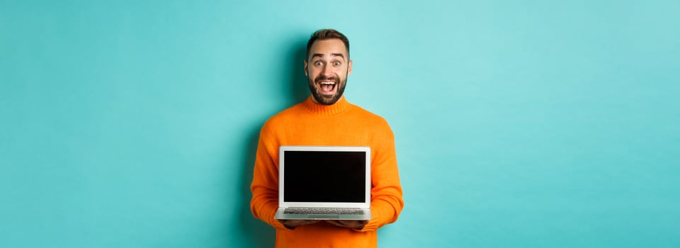 Handsome bearded man in orange sweater showing laptop screen, demonstrating promo, standing over light blue background photo