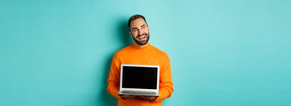 Happy caucasian man thinking and showing laptop screen, smiling pleased while pondering, standing in orange sweater against light blue background photo