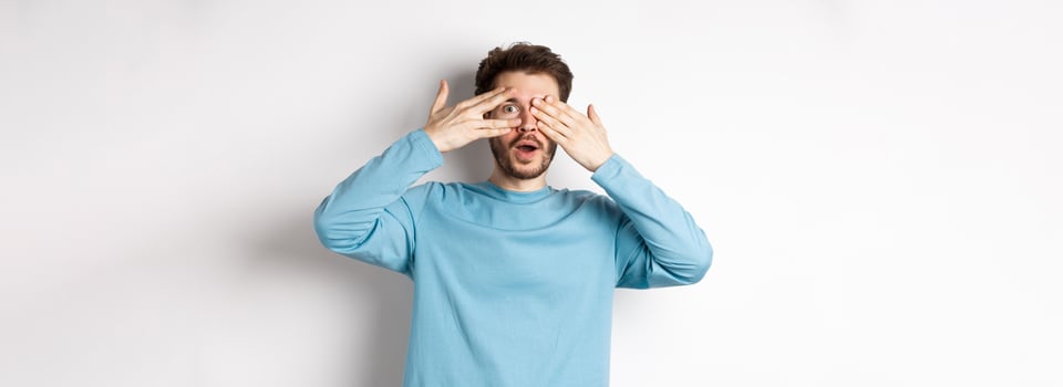 Amazed smiling man covering eyes with hands but peeking through fingers at something awesome, standing over white background photo