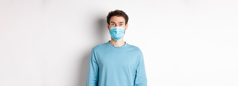 Covid-19, pandemic and social distancing concept. Cheerful man in medical mask raising eyebrows, looking surprised and excited at camera, standing over white background photo