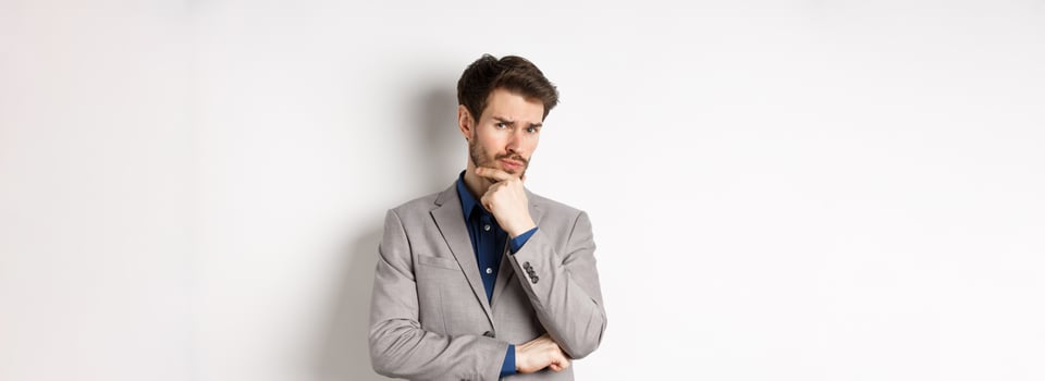 Hesitant young businessman in suit looking skeptical at camera, thinking and making decision, standing against white background photo