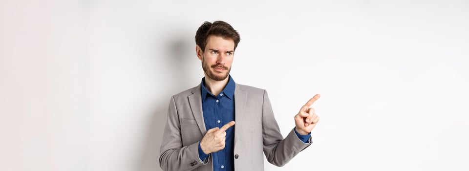Displeased businessman in suit pointing and looking left with skeptical smirk, dislike banner, disapprove promo offer, standing on white background photo