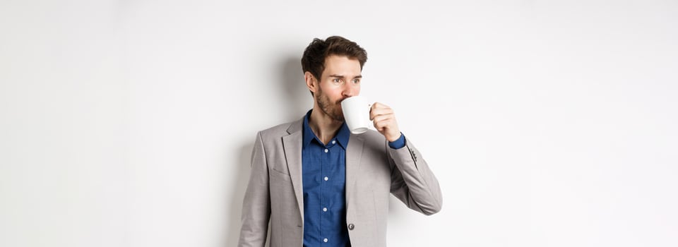 Handsome businessman in suit drinking coffee or tea from office mug, looking aside at logo, standing against white background photo
