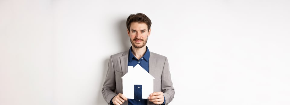 Real estate and insurance concept. Salesman in grey suit showing paper house cutout, selling property, smiling friendly at camera, white background photo