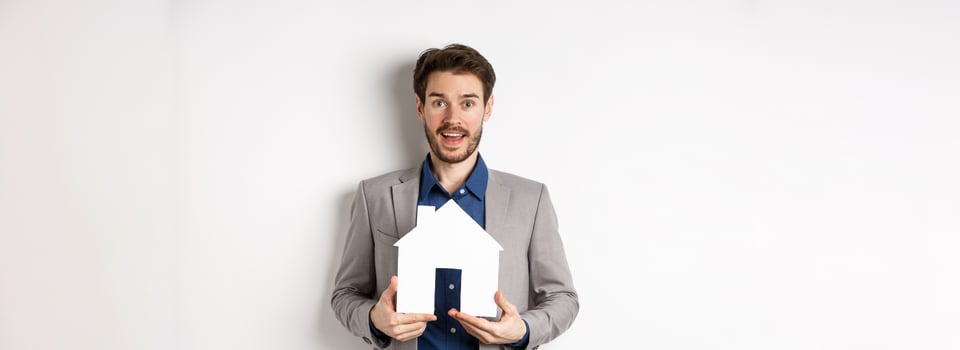 Real estate. Excited male entrepreneur holding paper house cutout, wearing suit and look hopeful at camera photo