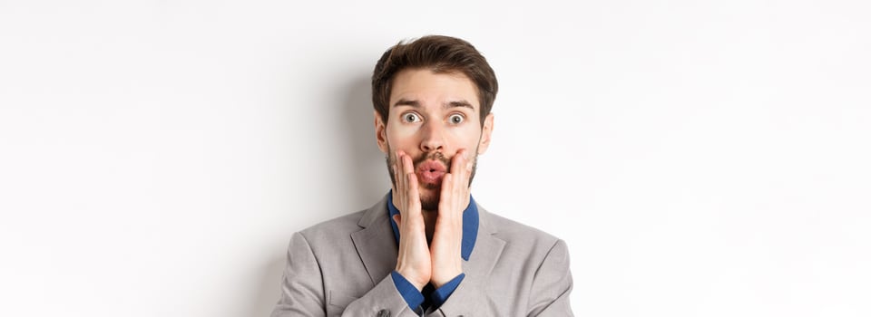 Surprised businessman in suit look with disbelied, pucker lips and touching face as staring at camera, standing on white background photo