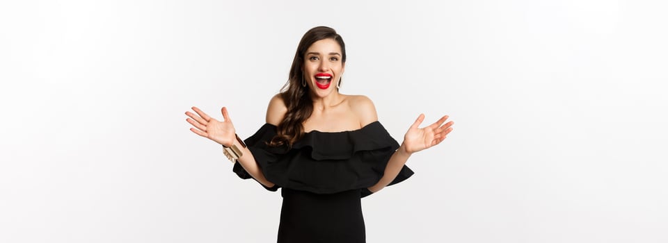 Beauty and fashion concept. Image of surprised and happy young woman in party dress reacting to good news, raising hands up and smiling amazed, white background photo