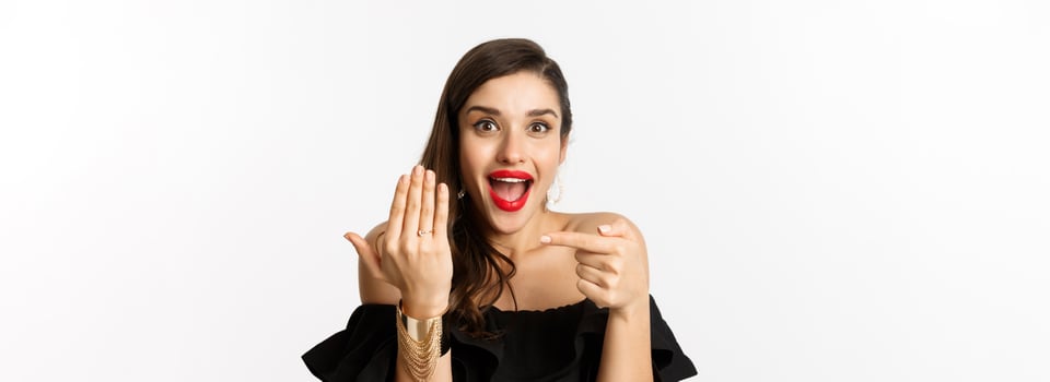 Happy young woman saying yes, become a bride, pointing at engagement ring on finger and smiling pleased, standing over white background photo