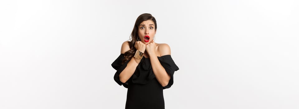 Portrait of glamour woman looking scared and shocked at camera, staring at something with fear, standing in black dress against white background photo