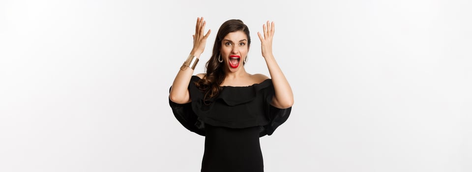 Fashion and beauty. Angry woman in black dress, shouting mad and shaking hands, grimacing outraged at camera, standing over white background photo