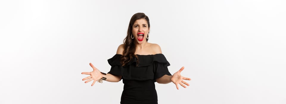 Fashion and beauty. Angry woman in black dress, shouting mad and shaking hands, grimacing outraged at camera, standing over white background photo