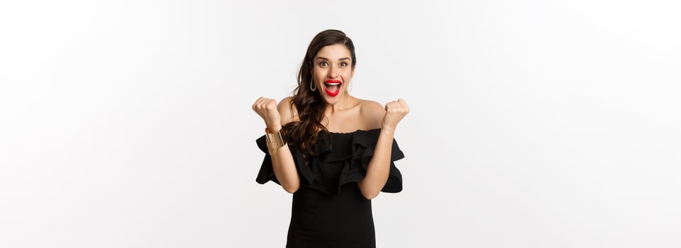 Fashion and beauty. Successful pretty woman in black dress celebrating, rejoicing of winning, triumphing over white background photo