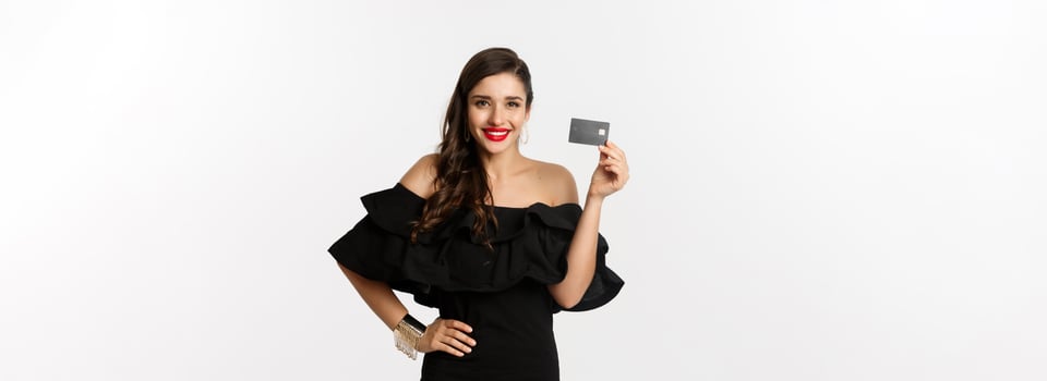 Beauty and shopping concept. Stylish pretty woman with red lips, black dress, smiling happy and showing credit card, standing over white background photo