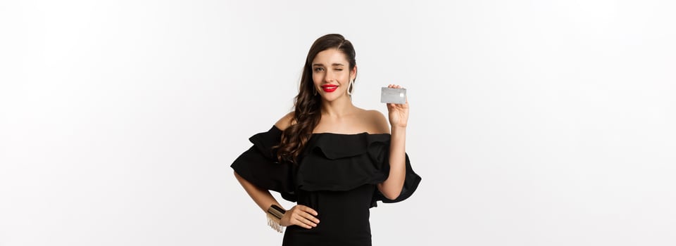 Beauty and shopping concept. Fashionable woman in black dress, wearing makeup, winking at camera and showing credit card, standing over white background photo