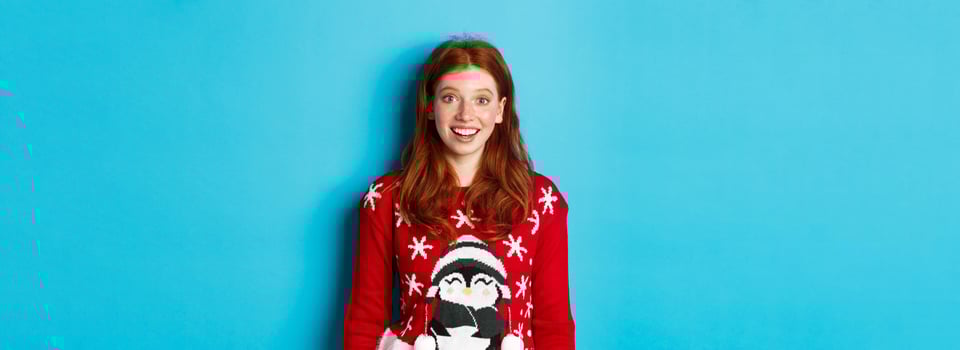 Winter holidays and Christmas Eve concept. Excited redhead girl in xmas sweater looking surprised at camera, standing against blue background photo