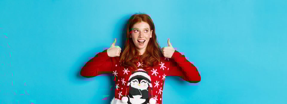 Winter holidays and Christmas Eve concept. Beautiful redhead girl in xmas sweater, celebrating New Year, showing thumbs up and looking at upper left corner, blue background photo