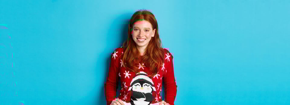 Winter holidays and Christmas Eve concept. Cute redhead girl wearing adorable sweater and smiling, celebrating New Year, standing over blue background photo