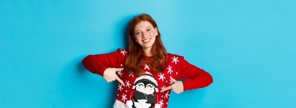 Winter holidays and Christmas Eve concept. Pretty redhead girl pointing fingers at cute xmas sweater with penguin, standing over blue background photo
