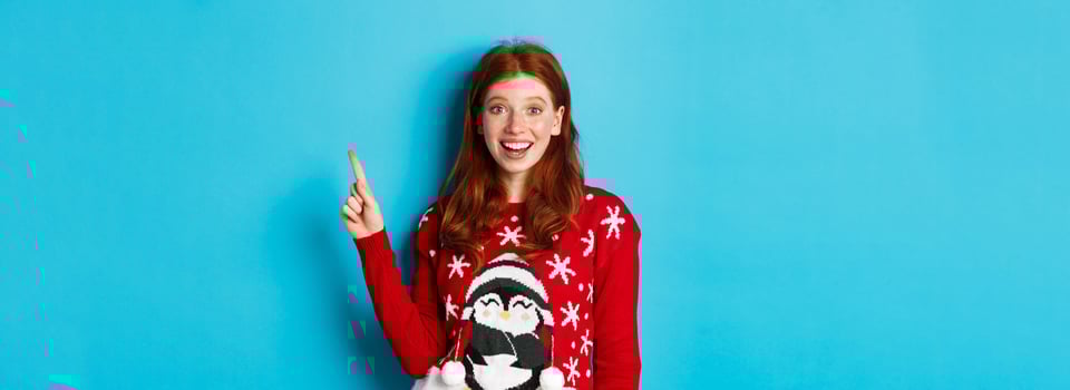 Merry Christmas. Cheerful redhead girl in xmas sweater, pointing finger at upper right corner and smiling excited, showing new year promo photo