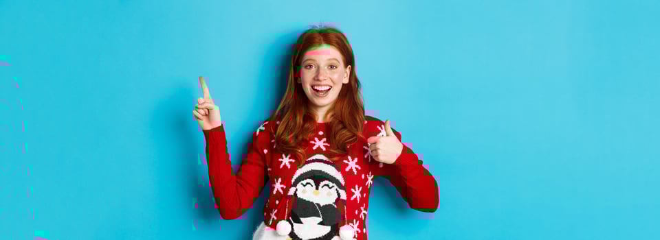 Merry Christmas. Cheerful redhead girl in xmas sweater, pointing finger at upper right corner, showing new year promo and thumbs-up in approval, praise product photo